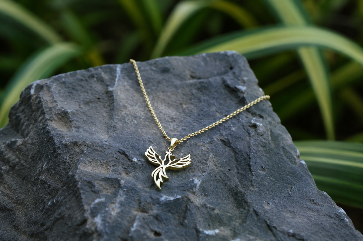 A gold pendant necklace featuring a stylized bird or phoenix design, resting on a dark textured rock with blurred green foliage in the background.