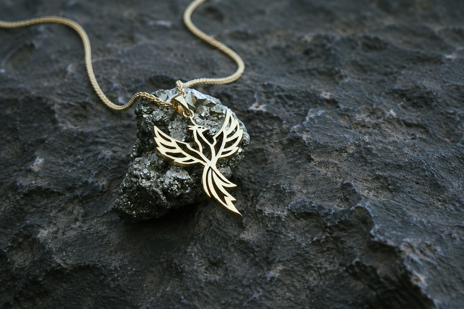 A gold toned pendant necklace with a stylized bird or flame-like cutout design, resting on a dark textured rock with a matching chain visible against a dark volcanic-looking surface.
