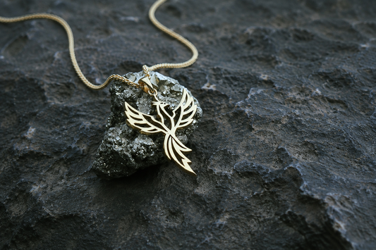 A gold toned pendant necklace with a stylized bird or flame-like cutout design, resting on a dark textured rock with a matching chain visible against a dark volcanic-looking surface.